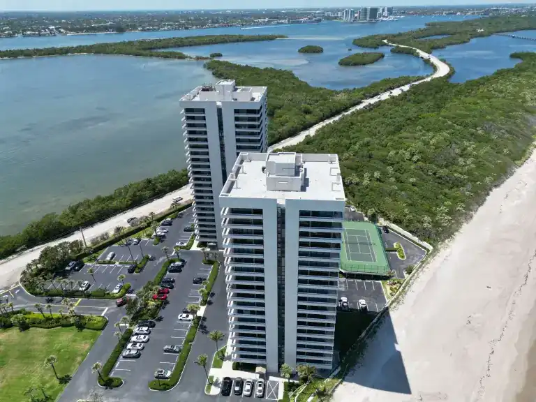 White flat roofs on twin beachfront towers with tennis courts below and a lagoon beyond in Riviera Beach, photographed April 2024