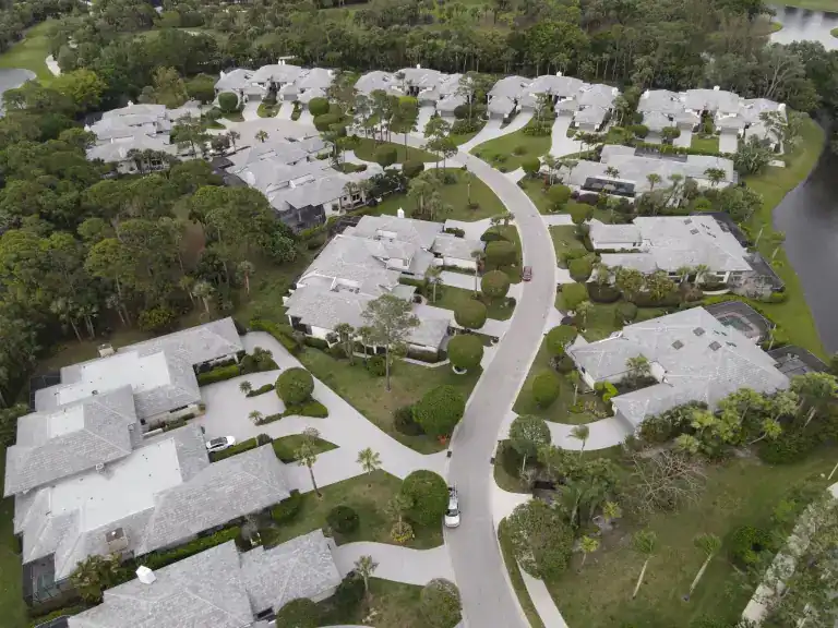 Wide aerial view of pale tile-roofed homes around a winding road between dense trees and a lake in West Palm Beach, photographed April 2024